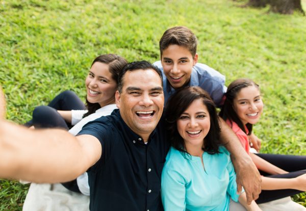 A happy latin family of five taking a selfie and smiling in a horizontal medium shot outdoors.