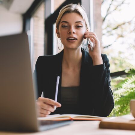 Portrait of smart casual Caucaisan female employee with blurred netbook looking at camera while making mobile calling, successful freelancer in jacket making phone conversation via application